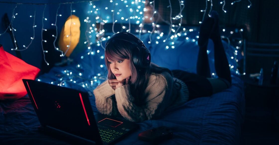 A young woman in a cozy room, wearing headphones and using a laptop, surrounded by warm fairy lights.